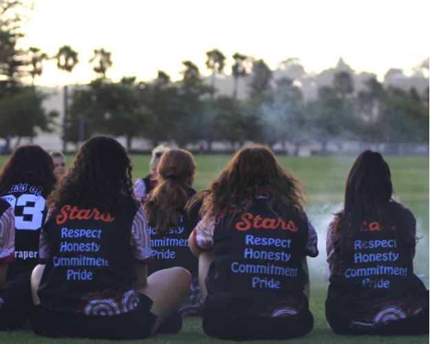 hree Aboriginal girls wearing Stars Foundation jackets sitting on grass, symbolizing future leadership and community engagement.